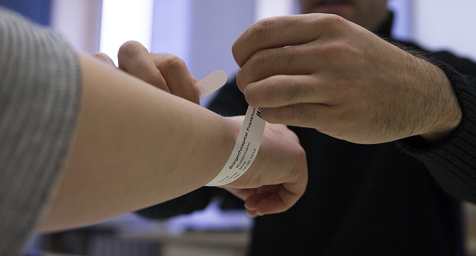 Nahaufnahme einer Hand, die einem Patienten ein Identifikationsarmband mit der Aufschrift "Bürgerhospital Frankfurt" anlegt