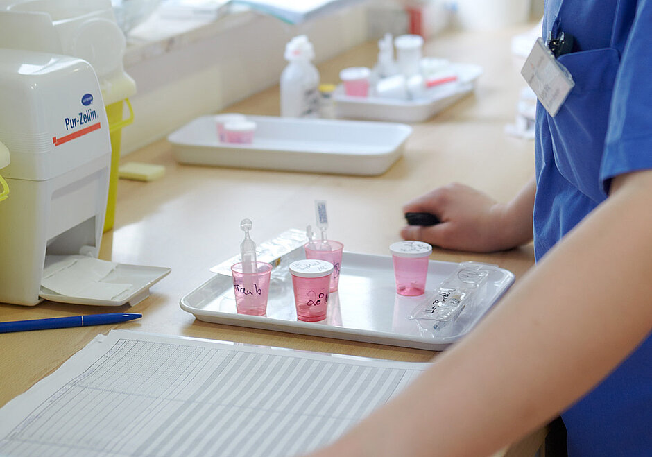 Ein Tablett mit vorbereiteten Medikamentendosen in kleinen rosa Bechern steht auf einer Arbeitsfläche. Eine Person in blauer medizinischer Kleidung ist im Hintergrund zu sehen, während sie eine Medikamentenvergabe durchführt. Die Becher sind beschriftet, und medizinische Utensilien liegen bereit.