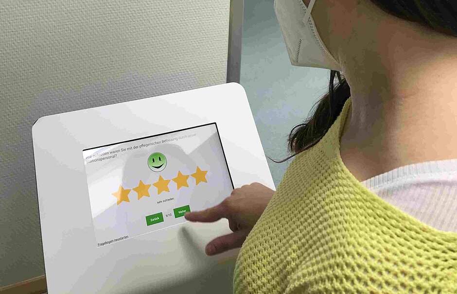 Eine Person mit Mundschutz gibt auf einem digitalen Bewertungsbildschirm im Bürgerhospital Frankfurt eine positive Rückmeldung zur pflegerischen Betreuung ab.
